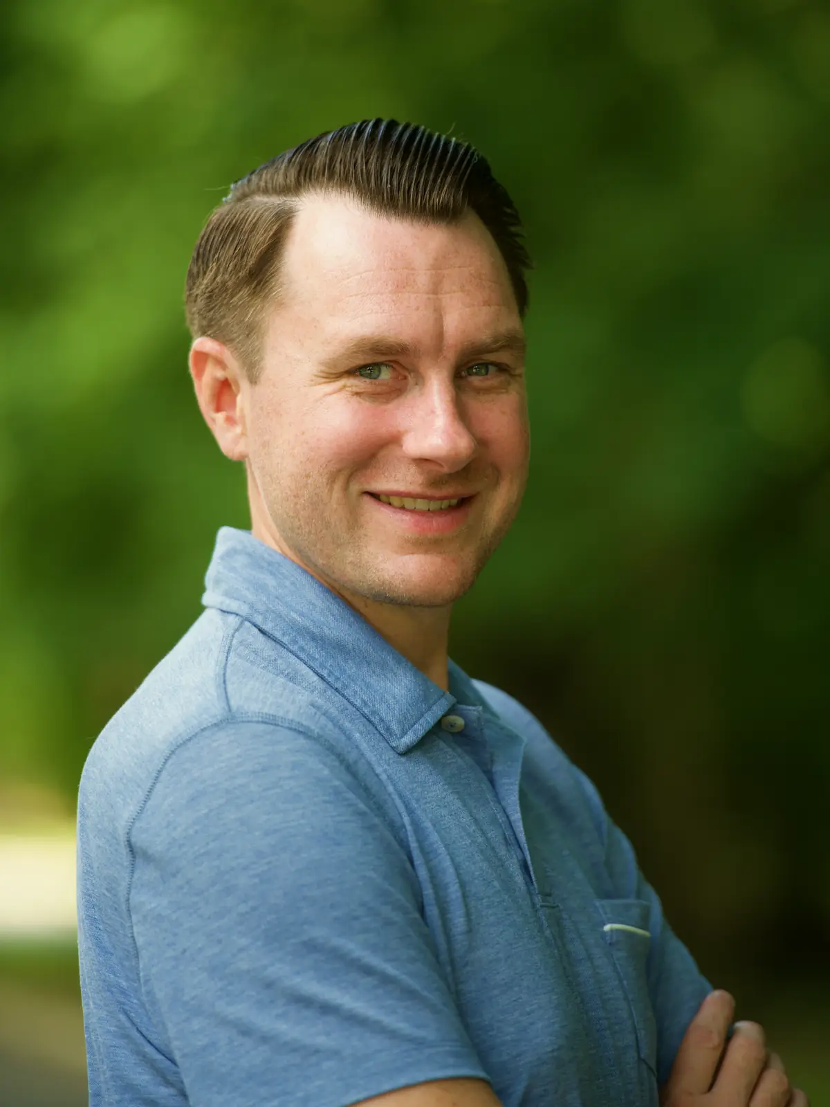 Portrait of Lee Procida, smiling with arms crossed, in front of green trees